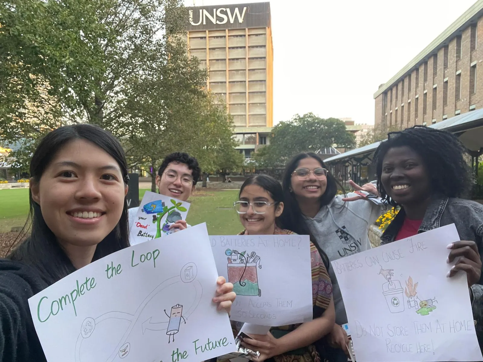 UNSW students holding hand-drawn battery and e-waste recycling posters on campus
